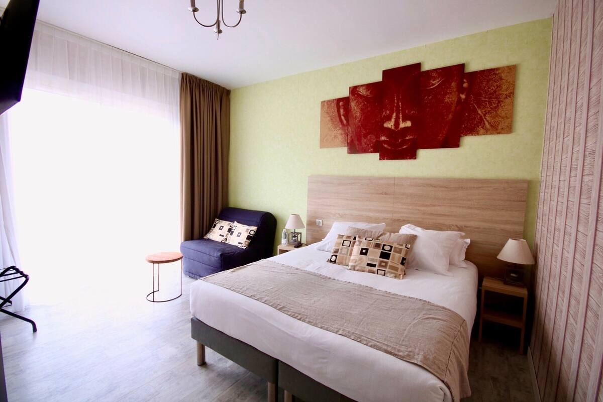 A hotel room with a bed, a chair, a nightstand, and a wall with artwork.
