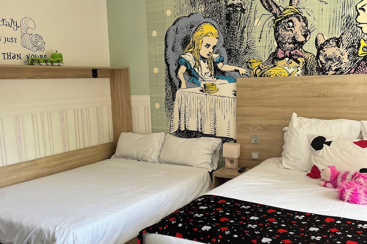 A hotel room with two single beds, a wooden headboard, a bedside table with a lamp, and a wall mural.
