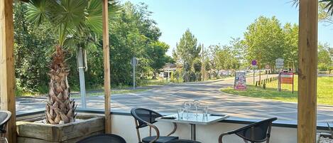 Une terrasse extérieure avec des tables et des chaises en bois, un palmier et une vue sur une route avec des panneaux de signalisation.