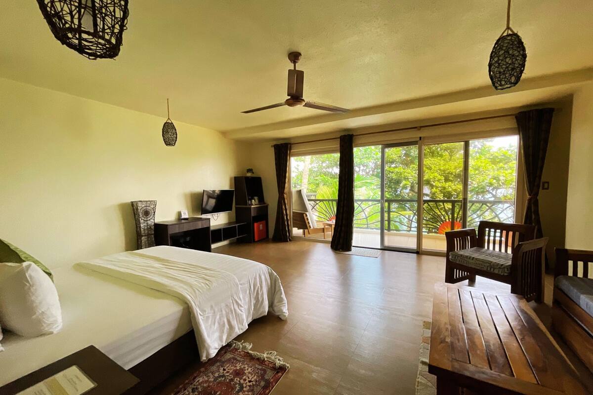 A spacious room with a bed, a wooden table, a TV, and a balcony with a view of greenery.