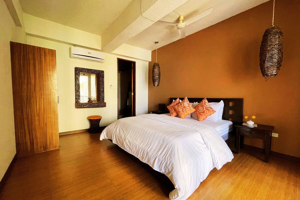A bedroom with a large bed, wooden flooring, a ceiling fan, a wall-mounted air conditioner, and a hanging decorative item.