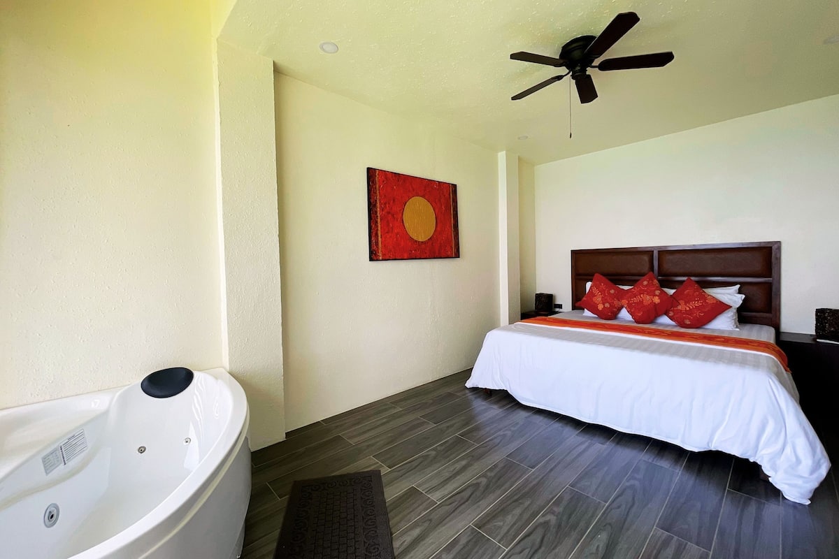 A hotel room with a large bathtub, a bed with red pillows, and a ceiling fan.