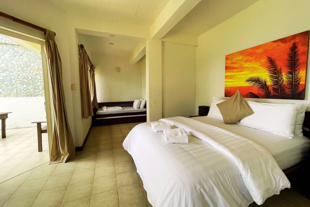 A hotel room with a large bed, a painting of a sunset, a balcony with curtains, and a stone wall.