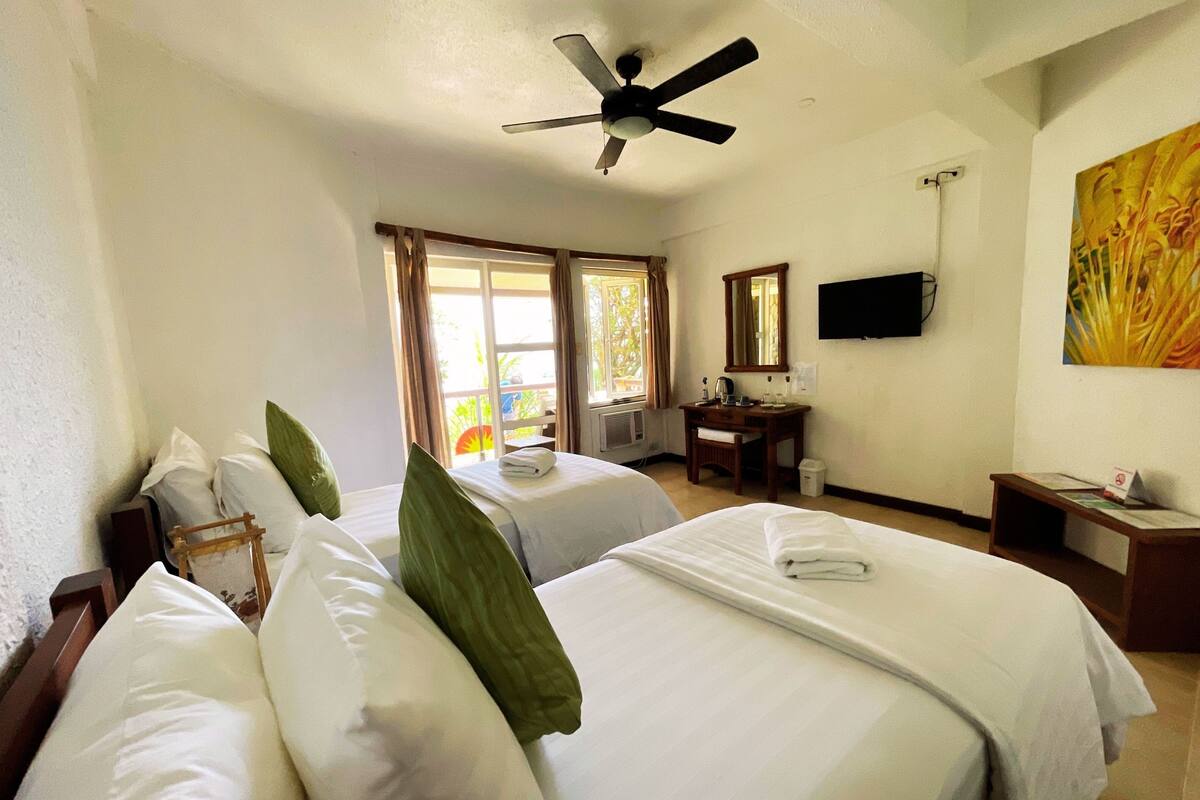 A hotel room with two beds, a ceiling fan, a desk with a chair, a television, and a window with curtains.