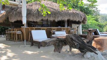 A beachside hut with thatched roof, wooden furniture, and a sandy ground.
