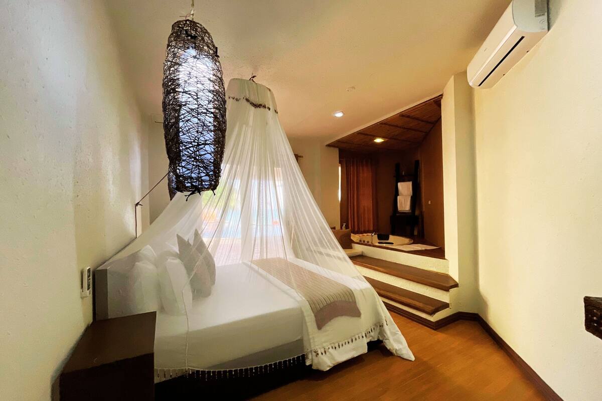 A bedroom with a bed under a mosquito net, a wooden staircase, and an air conditioning unit on the wall.