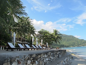 Eine Strandlounge mit Steinmauern und weißen Sonnenschirmen mit Blick auf einen Strand und eine gebirgige Landschaft.