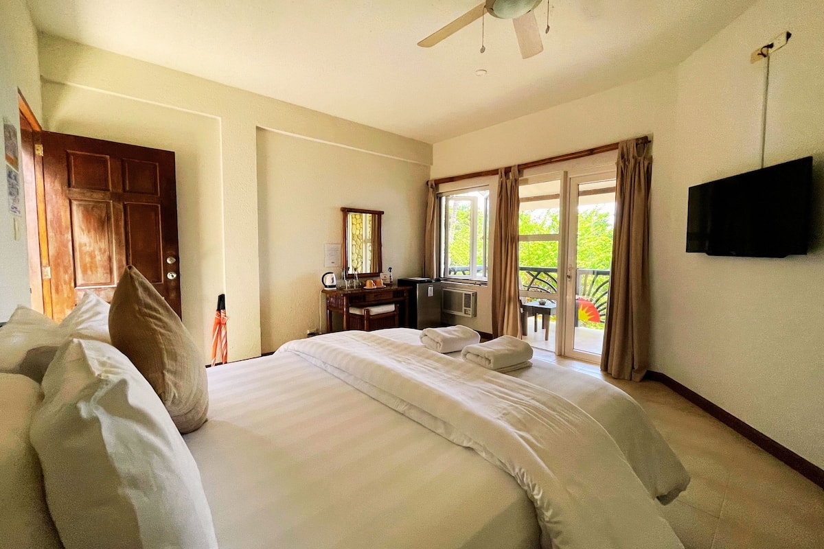 A hotel room with a large bed, a desk with a computer, a TV mounted on the wall, and a balcony with curtains.