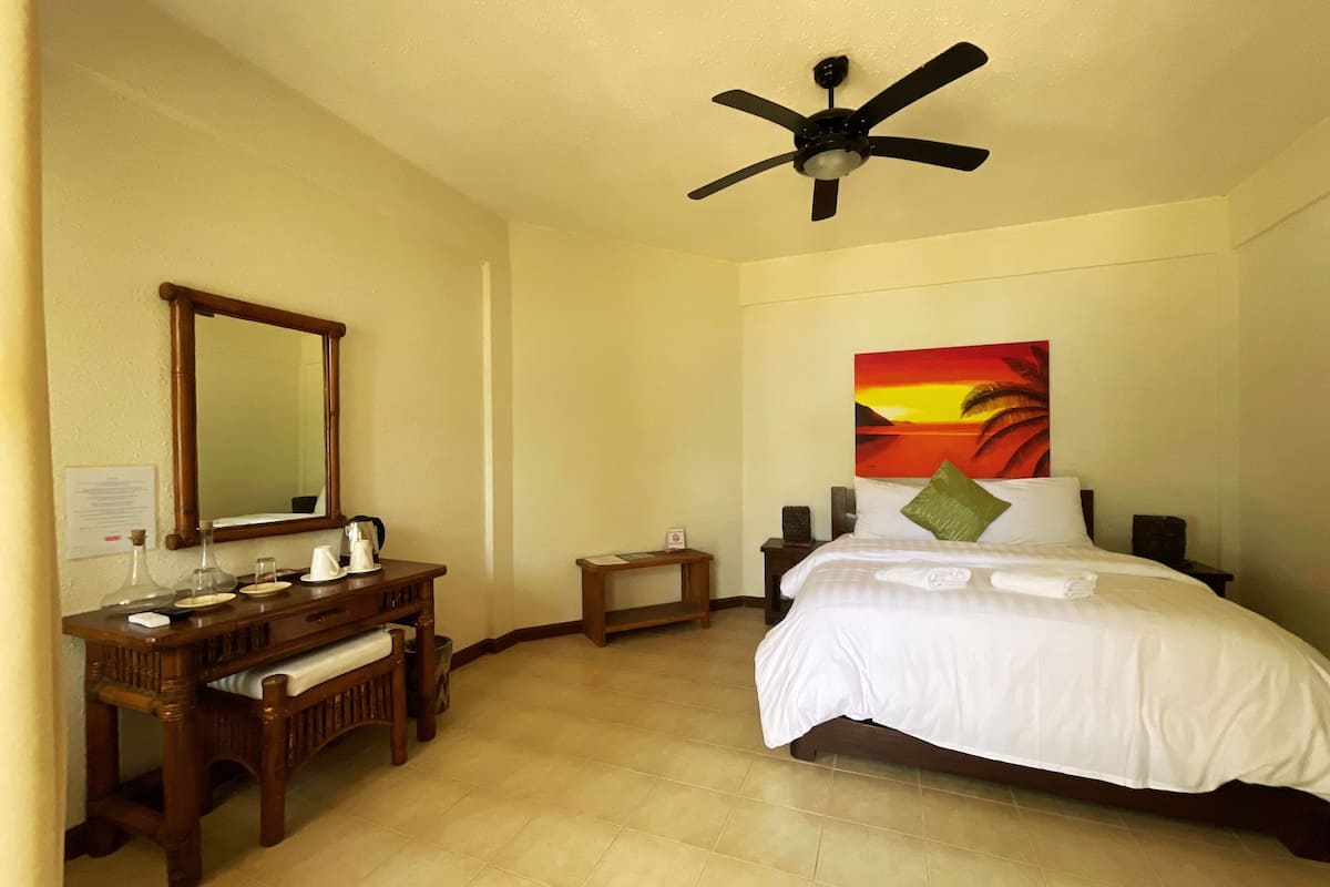 A hotel room with a bed, a wooden dresser, a ceiling fan, a painting, and a small table.