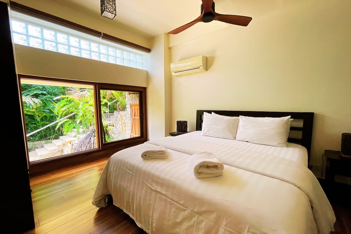A hotel room with a large bed, a ceiling fan, an air conditioning unit, and a view of greenery through a large window.