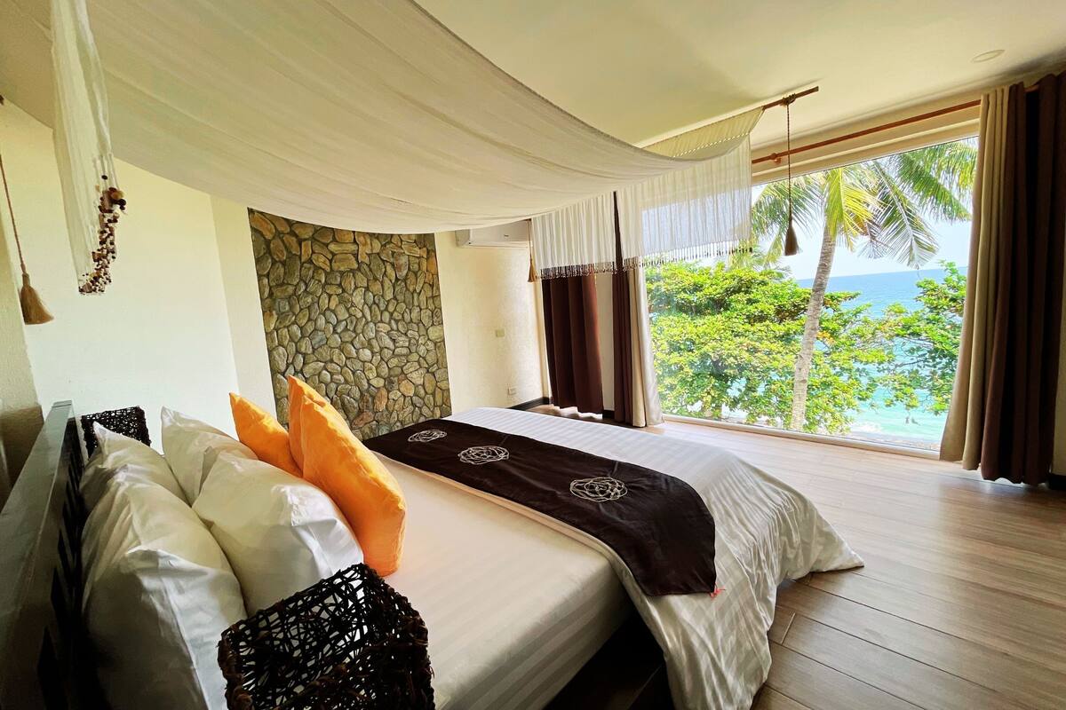 A bedroom with a large bed, a stone wall, and a view of the ocean.