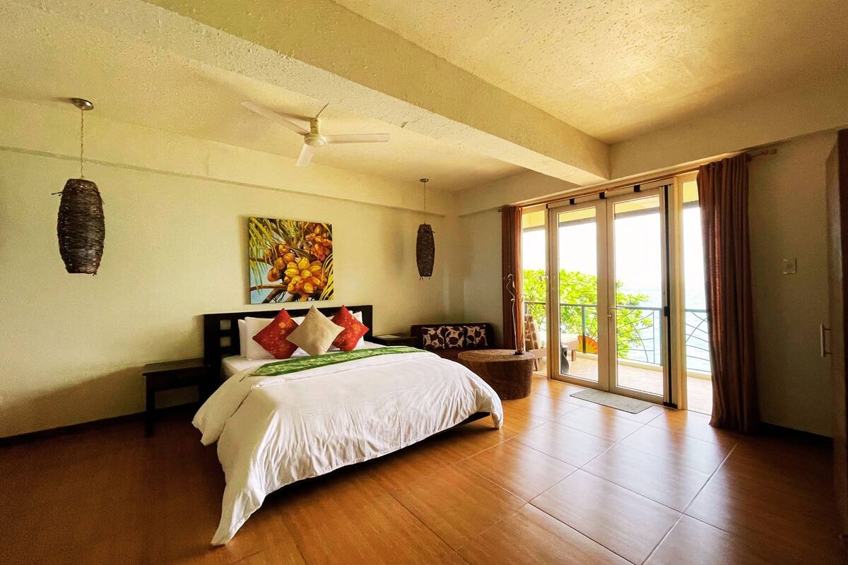 A spacious bedroom with a large bed, a ceiling fan, a painting, and a sliding glass door to a balcony.