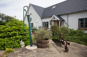 Una casa con techo inclinado, un jardín con manguera verde, un barril oxidado y una carretilla.