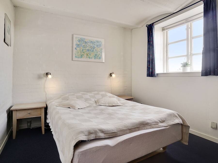 Un dormitorio con una cama, dos mesitas de noche, una lámpara de pared, una ventana con cortinas y un cuadro enmarcado en la pared.