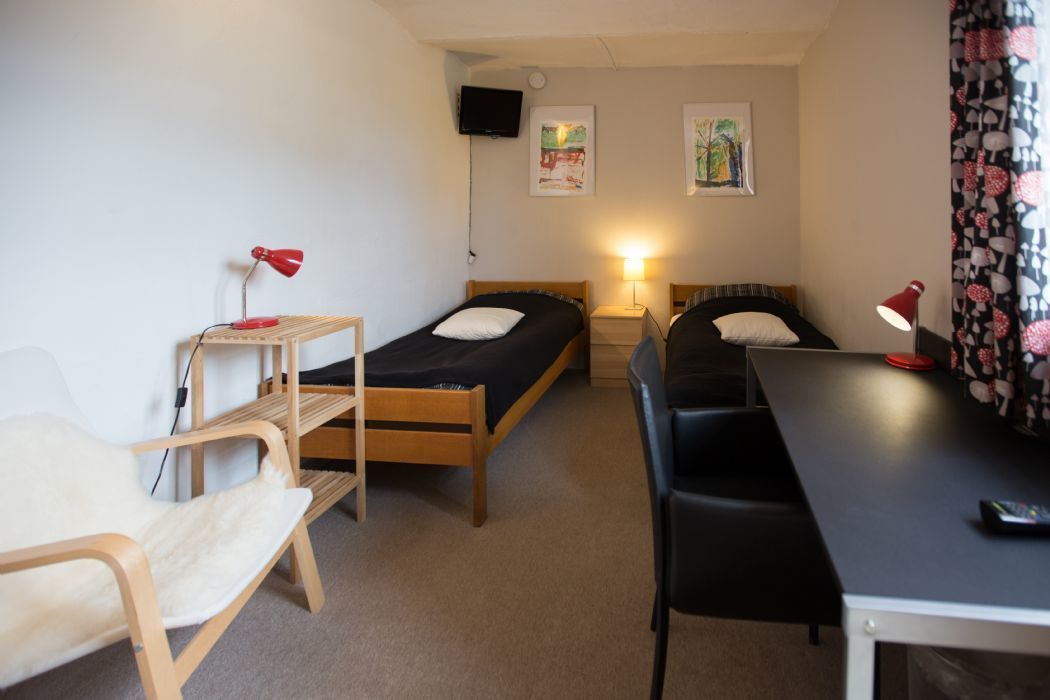 A small, well-lit room with two single beds, a desk, a chair, and a TV mounted on the wall.
