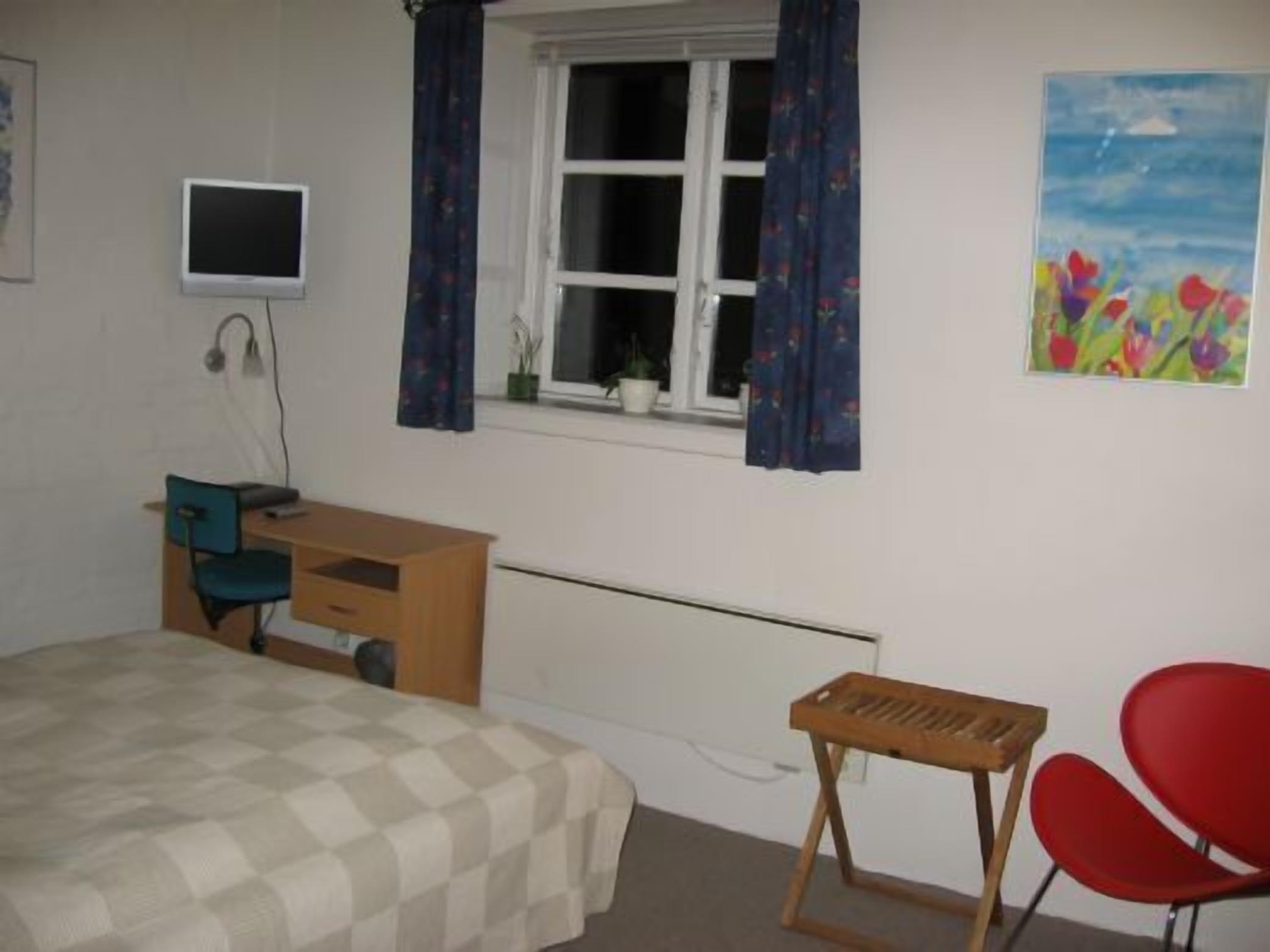 A bedroom with a bed, a desk, a chair, a window with curtains, a television, and a painting.