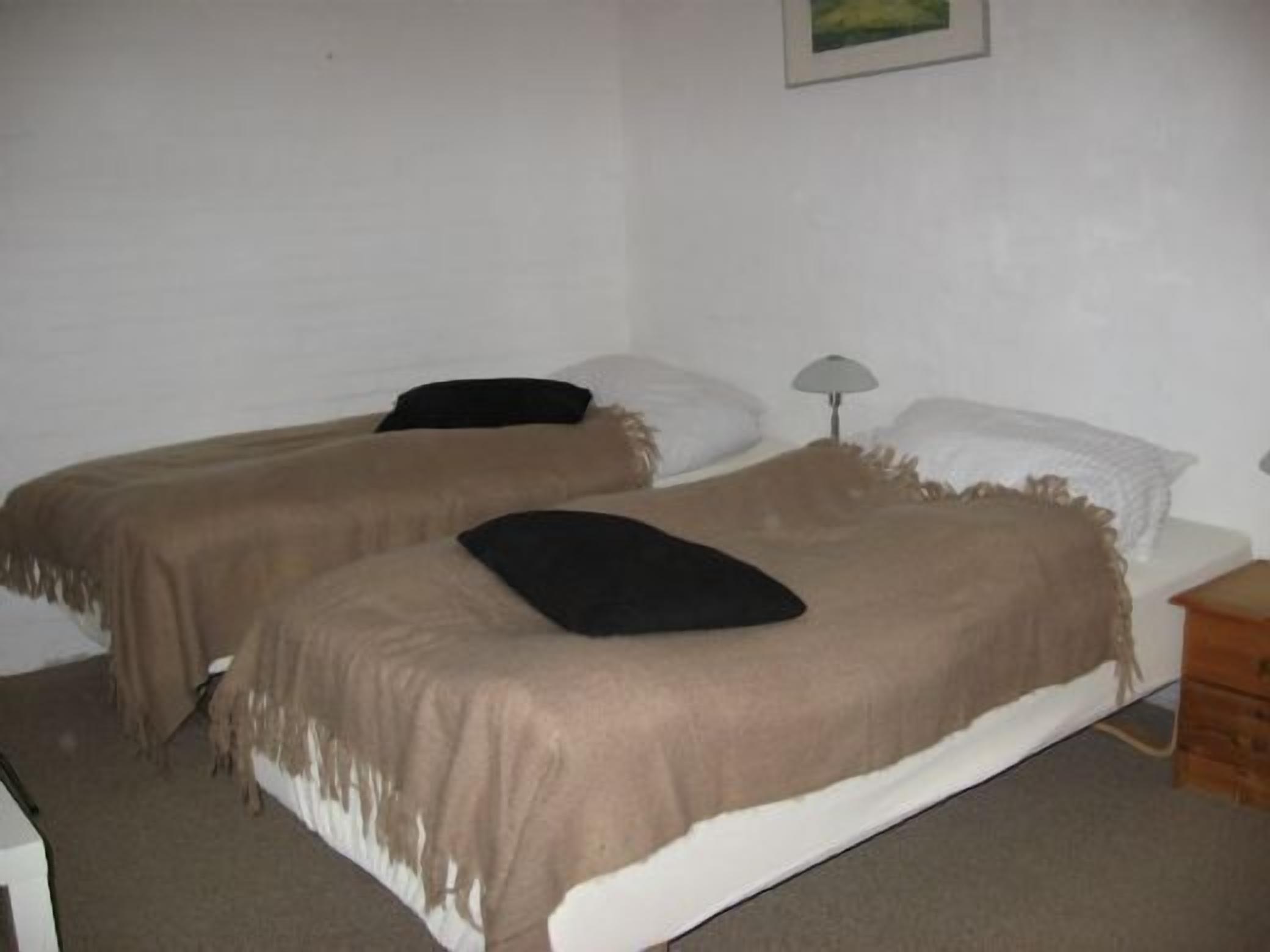 A simple room with two beds, each with a pillow and a blanket. There is a small bedside table with a lamp on one side.