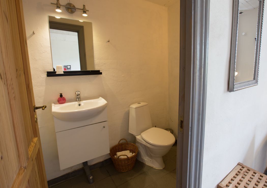 A small bathroom with a white sink, mirror, and toilet. There is a basket under the sink and a wooden door frame.