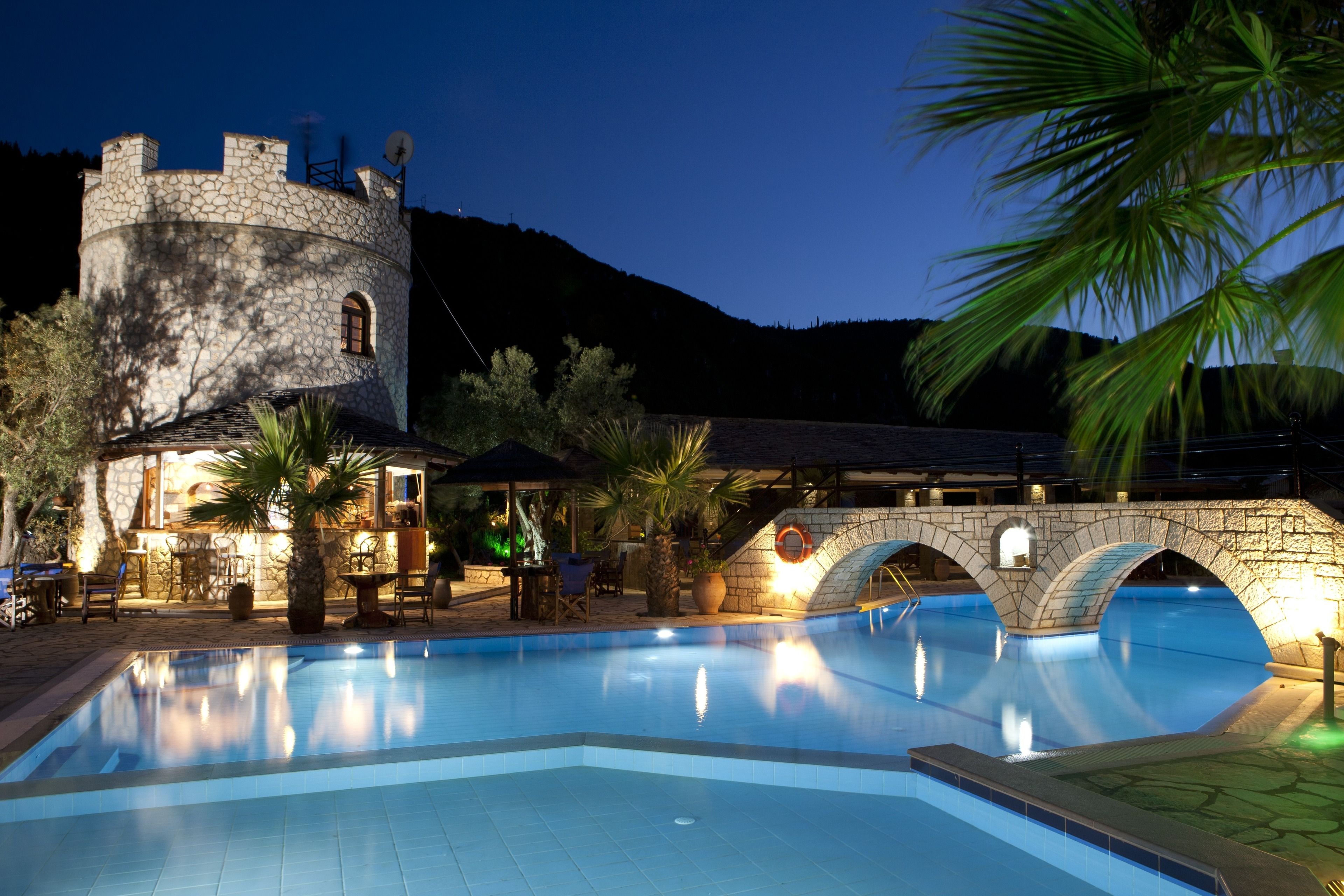 A swimming pool with stone architecture, a tower, and palm trees.