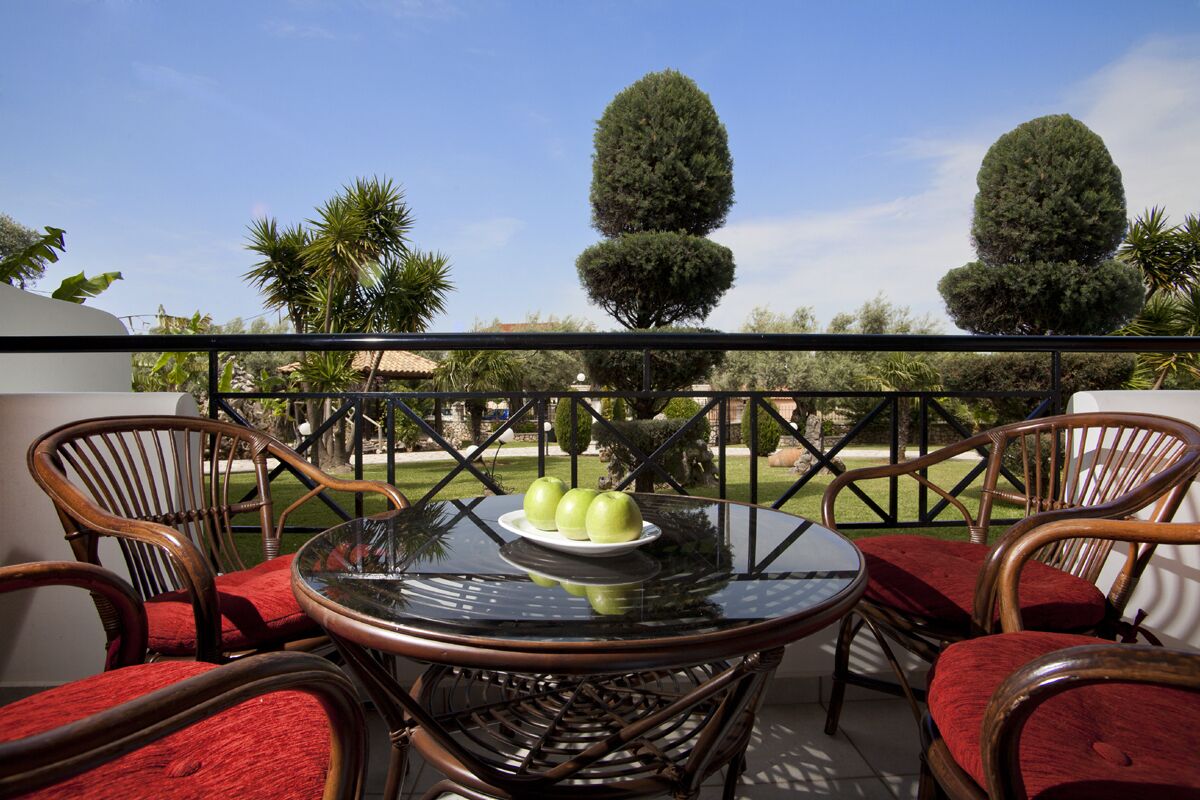 Un balcone con sedie in vimini e un tavolo in vetro, con un piatto di mele e vista su alberi curati.