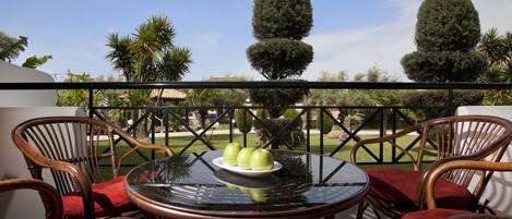 A balcony with wicker chairs and a glass table, featuring a plate of apples and a view of neatly trimmed trees.