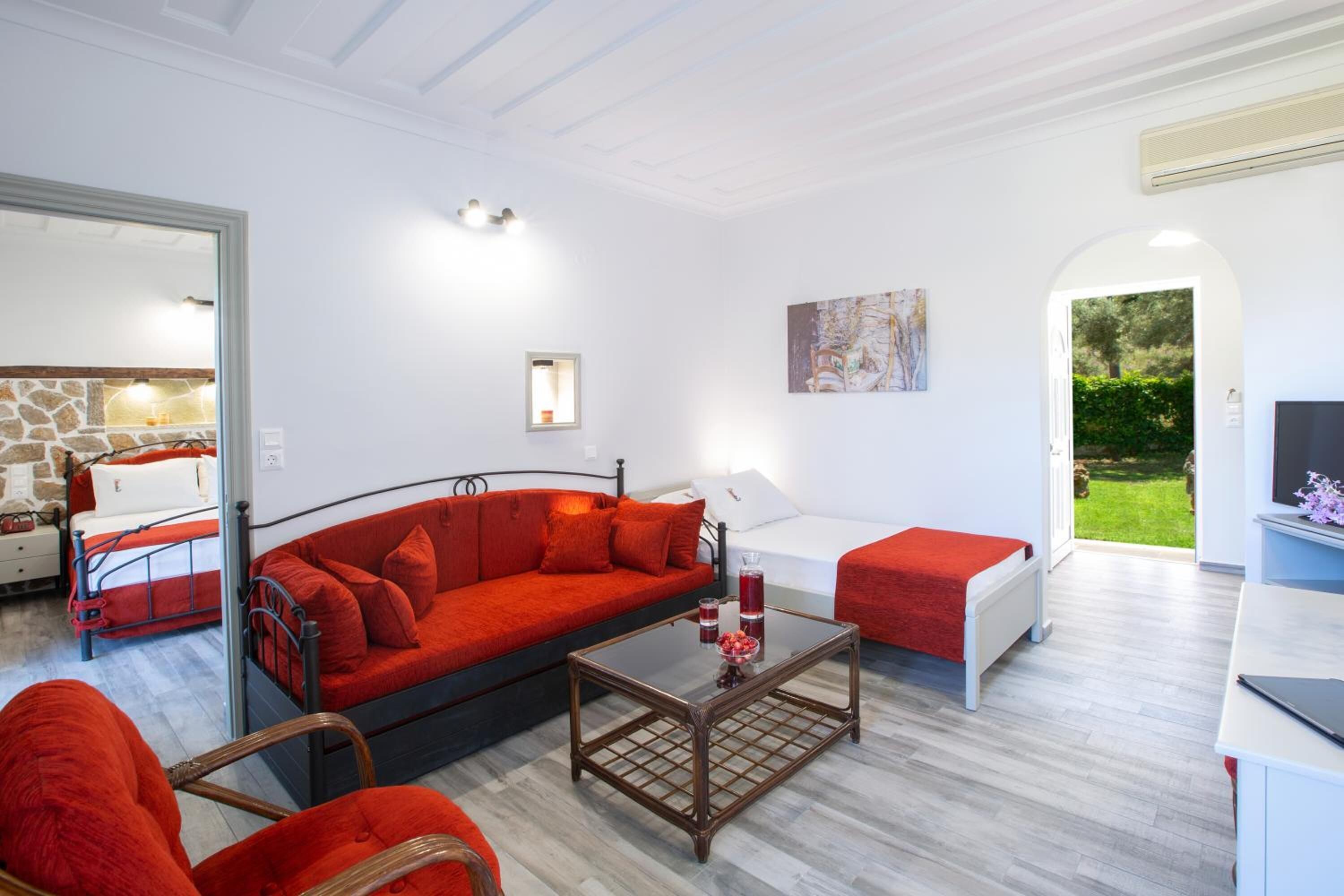 A modern living room with a red sofa, a glass coffee table, and a white armchair. There is a small table with a lamp and a painting on the wall.