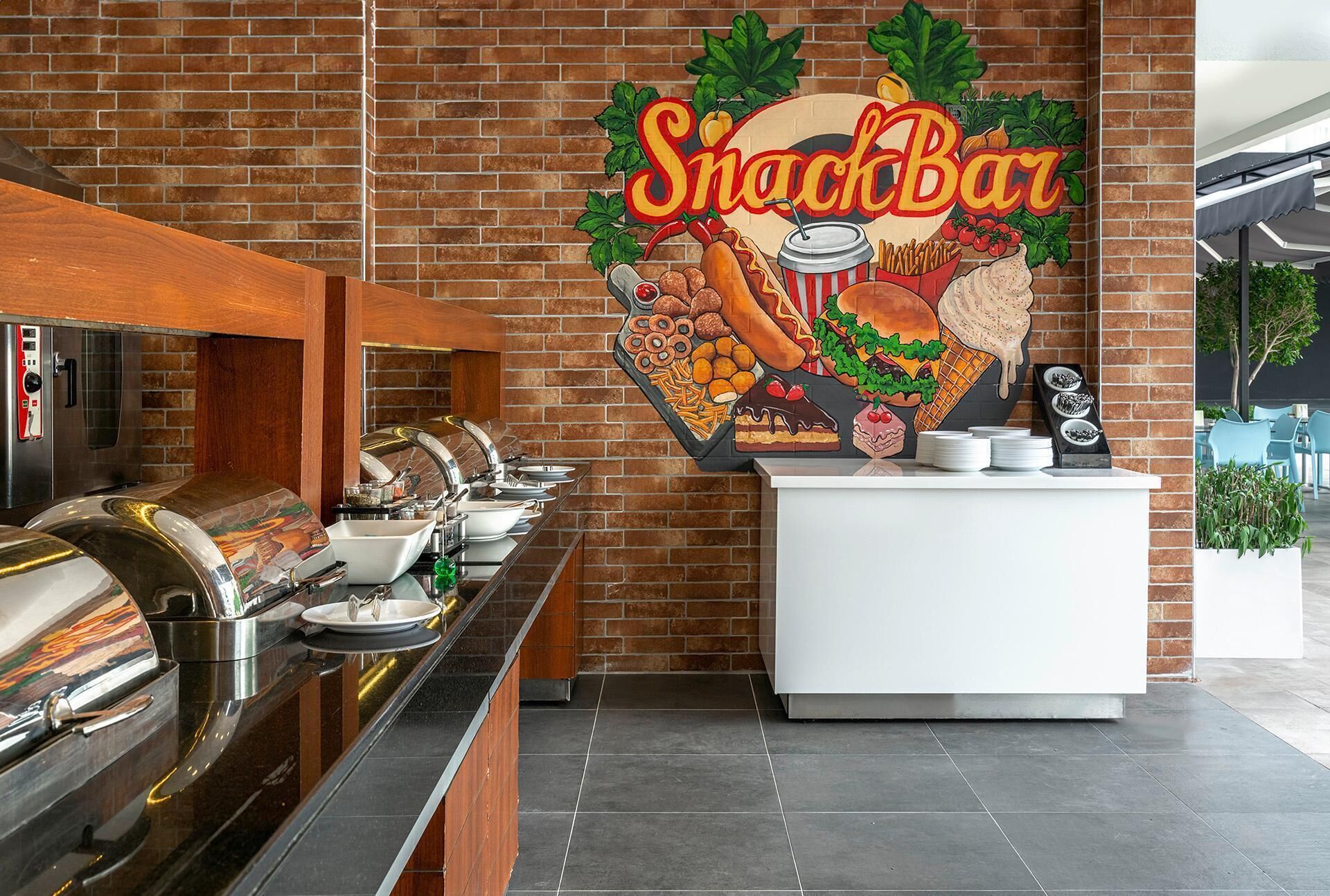Um bar de lanches com parede de tijolos ao fundo, um grande mural de comidas e um balcão de buffet com várias estações de serviço.