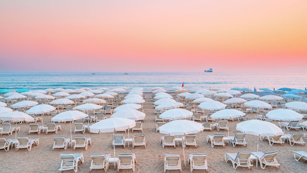 A beach with rows of white sun loungers and umbrellas, a calm sea, and a sunset in the background. - Avena Resort & Spa Hotel - All Inclusive  (Alanya)