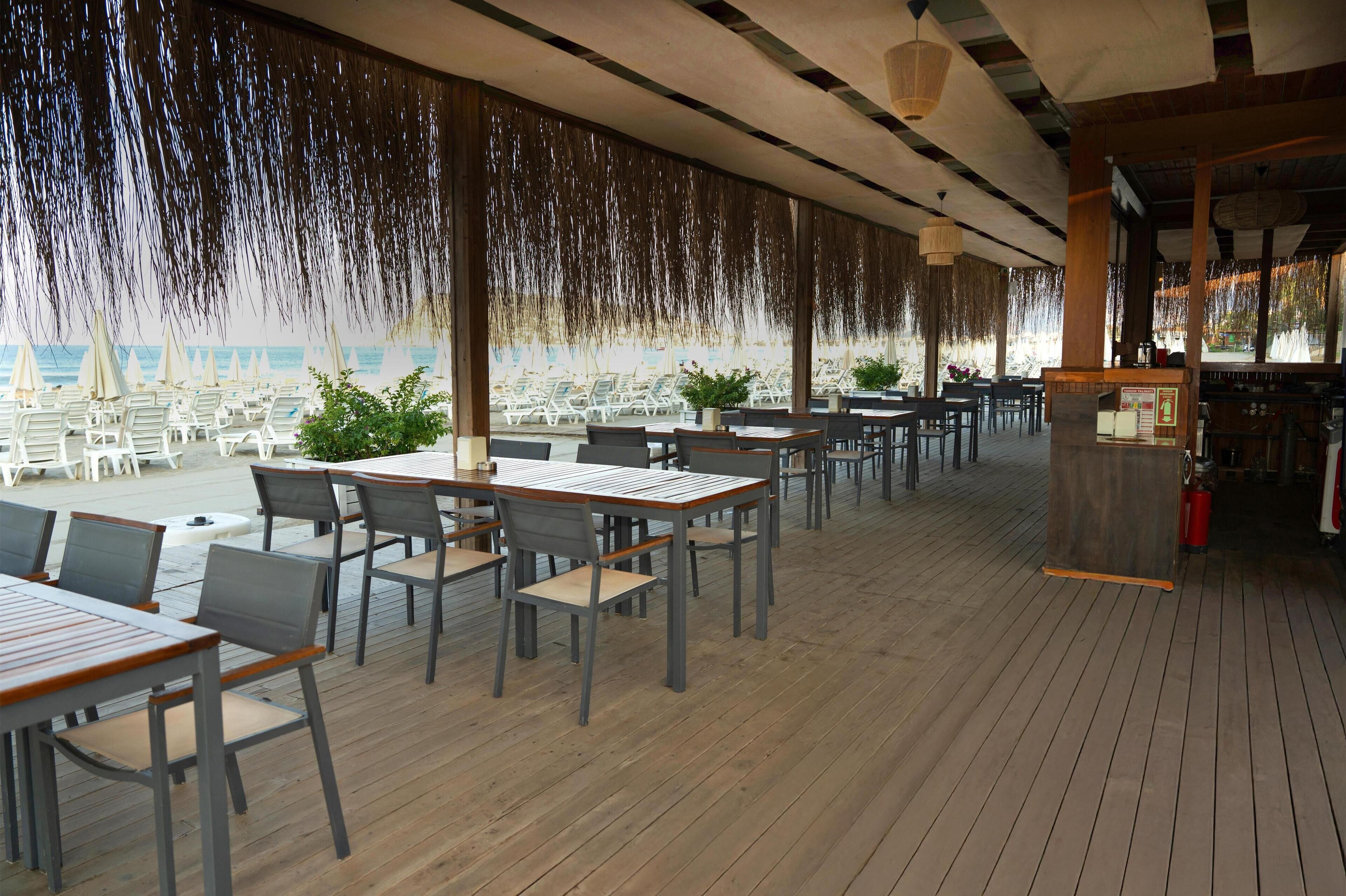 A covered outdoor dining area with tables and chairs, potted plants, and a view of the ocean.