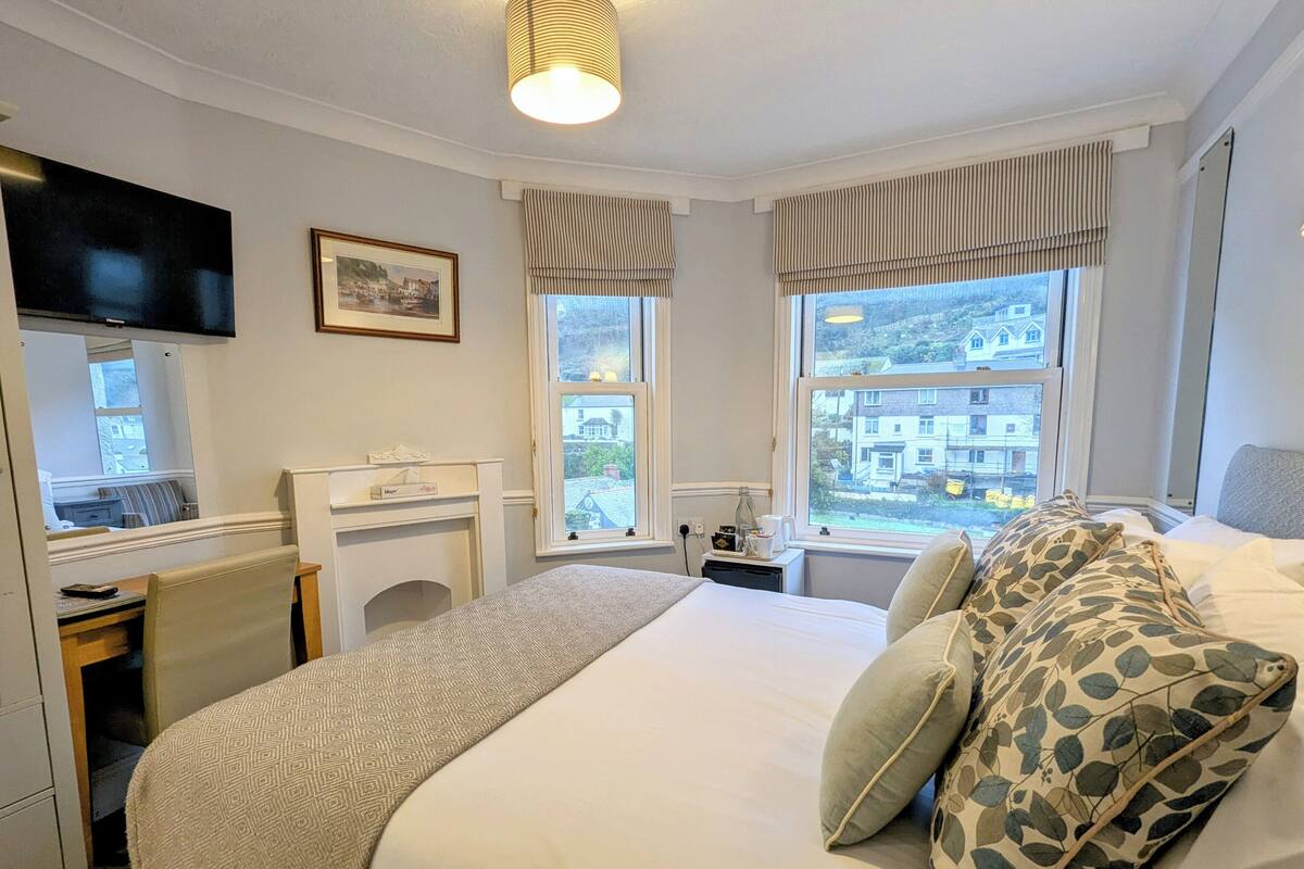 Ein Hotelzimmer mit Bett, Schreibtisch, Fernseher und einem Fenster mit Blick auf Gebäude.