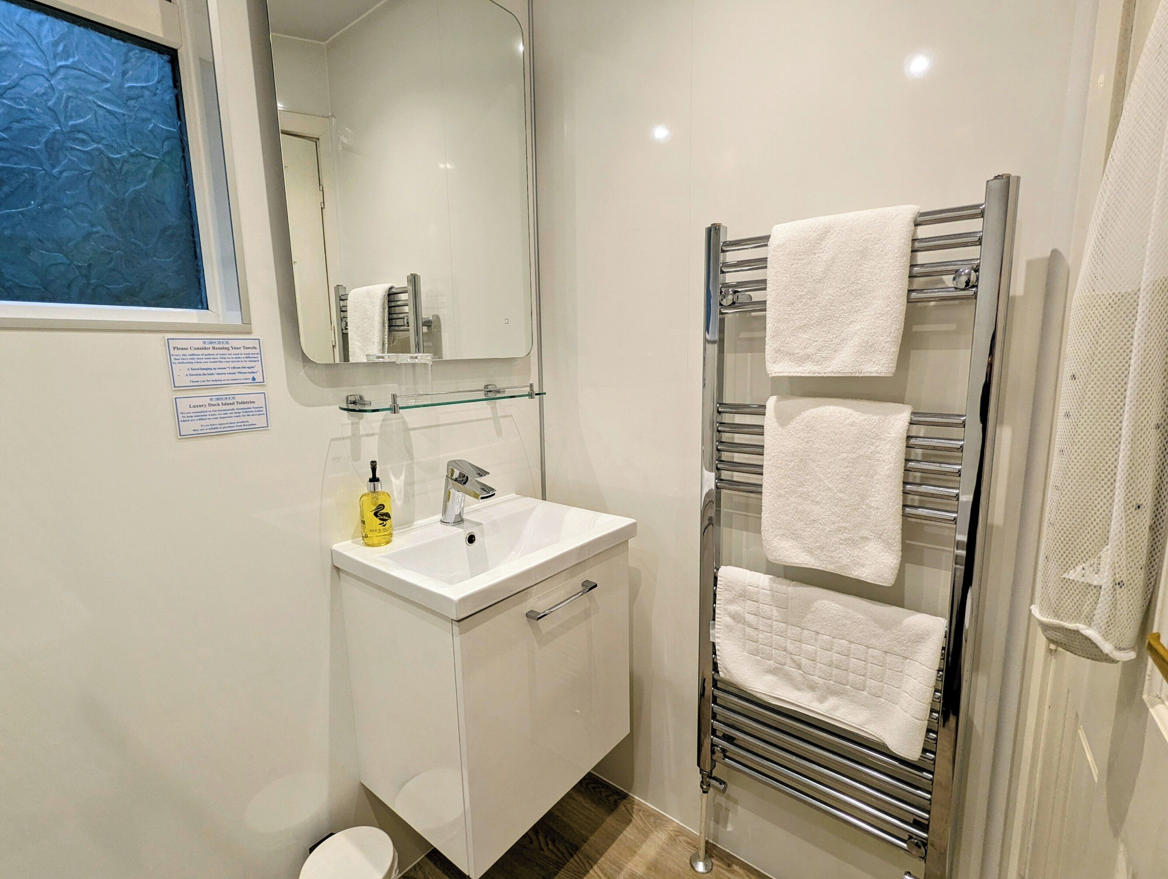 A modern bathroom with a white sink, a glass shelf, a towel rack with multiple towels, and a mirror above the sink.