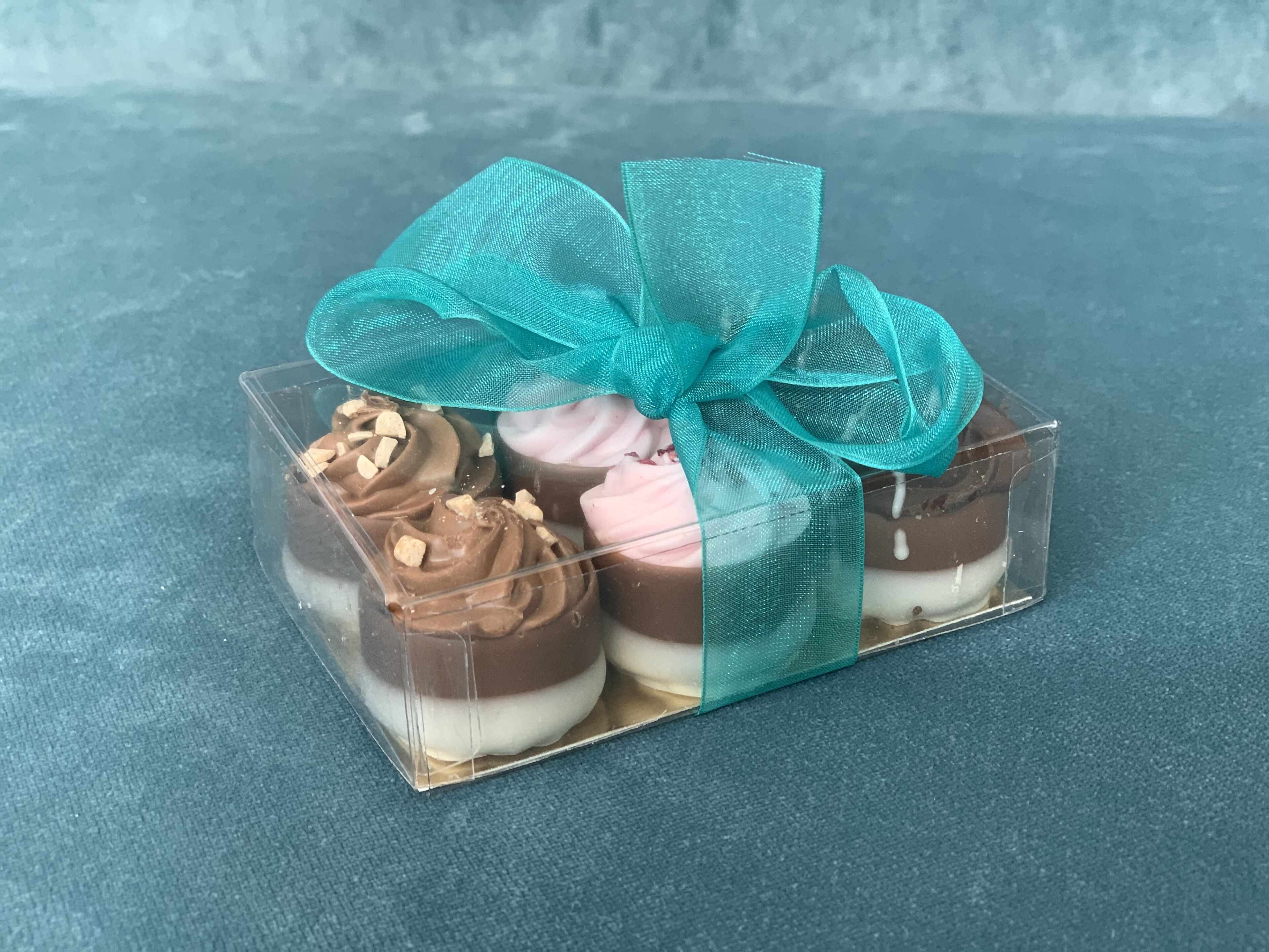 Una caja de plástico transparente con tres postres, cada uno coronado con un glaseado de diferente color y una cinta azul aguamarina.