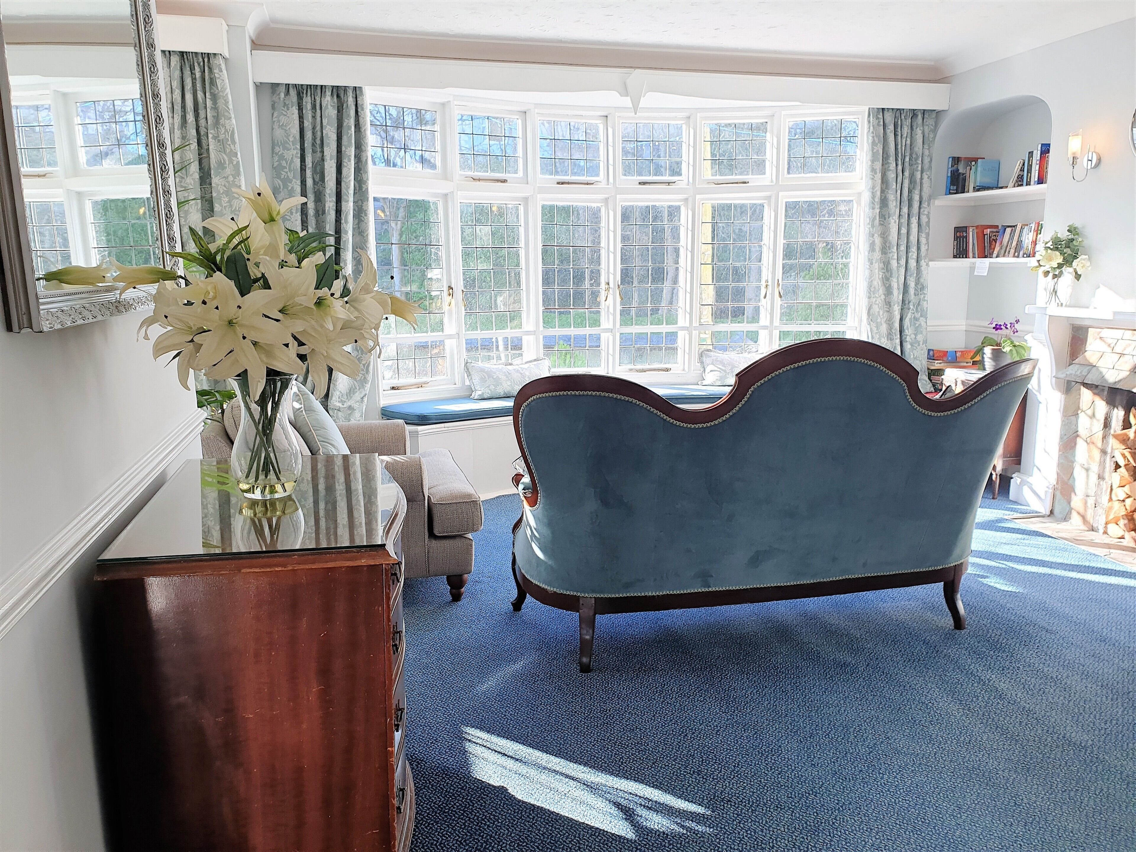 A room with a blue sofa, a wooden side table with a vase of flowers, a bookshelf, and a large window with curtains.