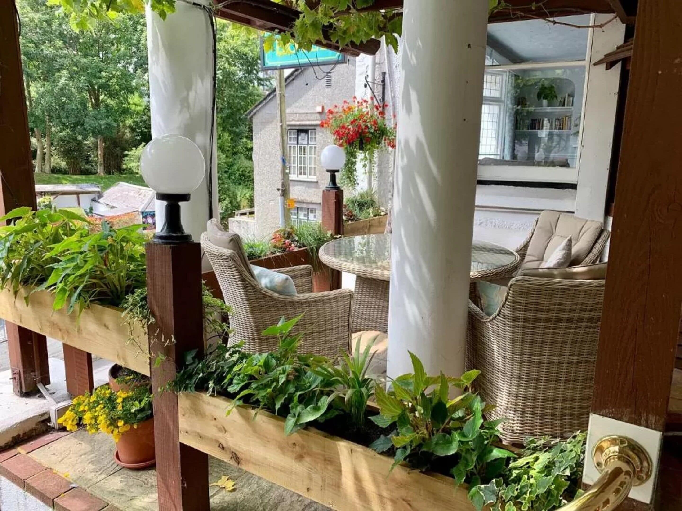 Een overdekt terras met rieten meubilair, een houten bank met planten en een glazen tafel.