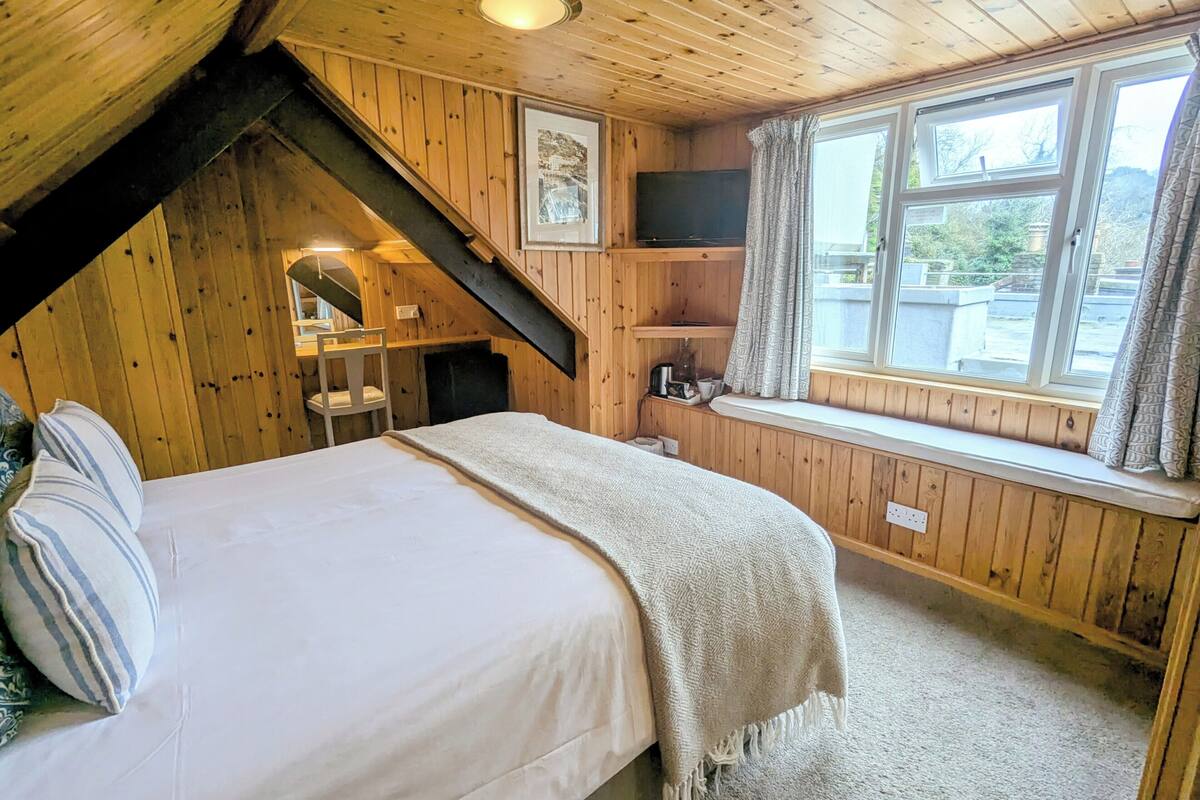 Ein gemütliches Schlafzimmer einer Holzhütte mit einem Bett, einem Fenster mit Vorhängen, einem an der Wand montierten Fernseher und einer kleinen Ablage mit Gegenständen.