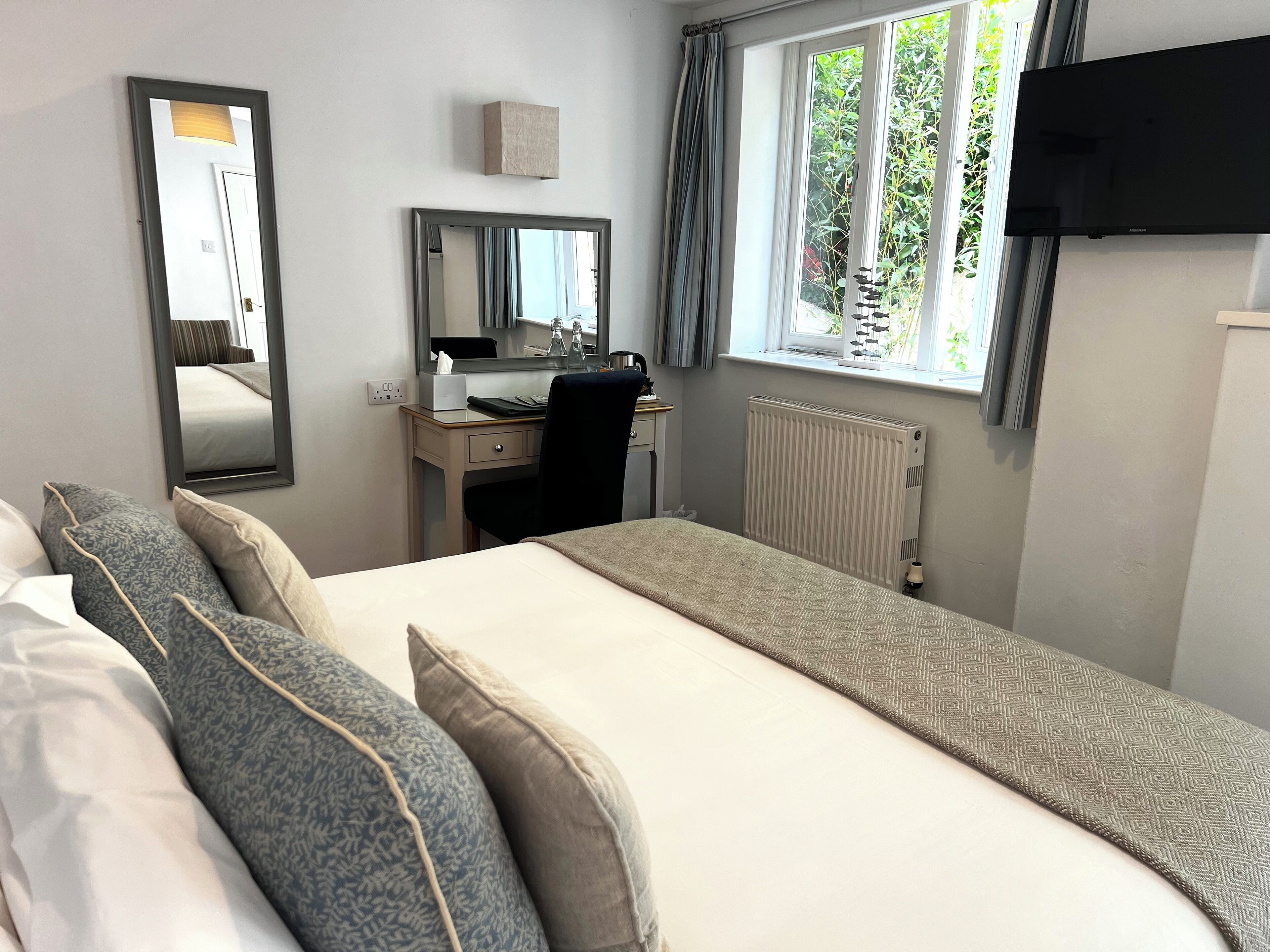 A hotel room with a bed, a desk, a chair, a mirror, a radiator, and a TV.