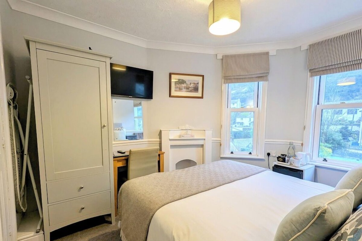 A bedroom with a bed, a desk, a chair, a wardrobe, a television, and a window with curtains.