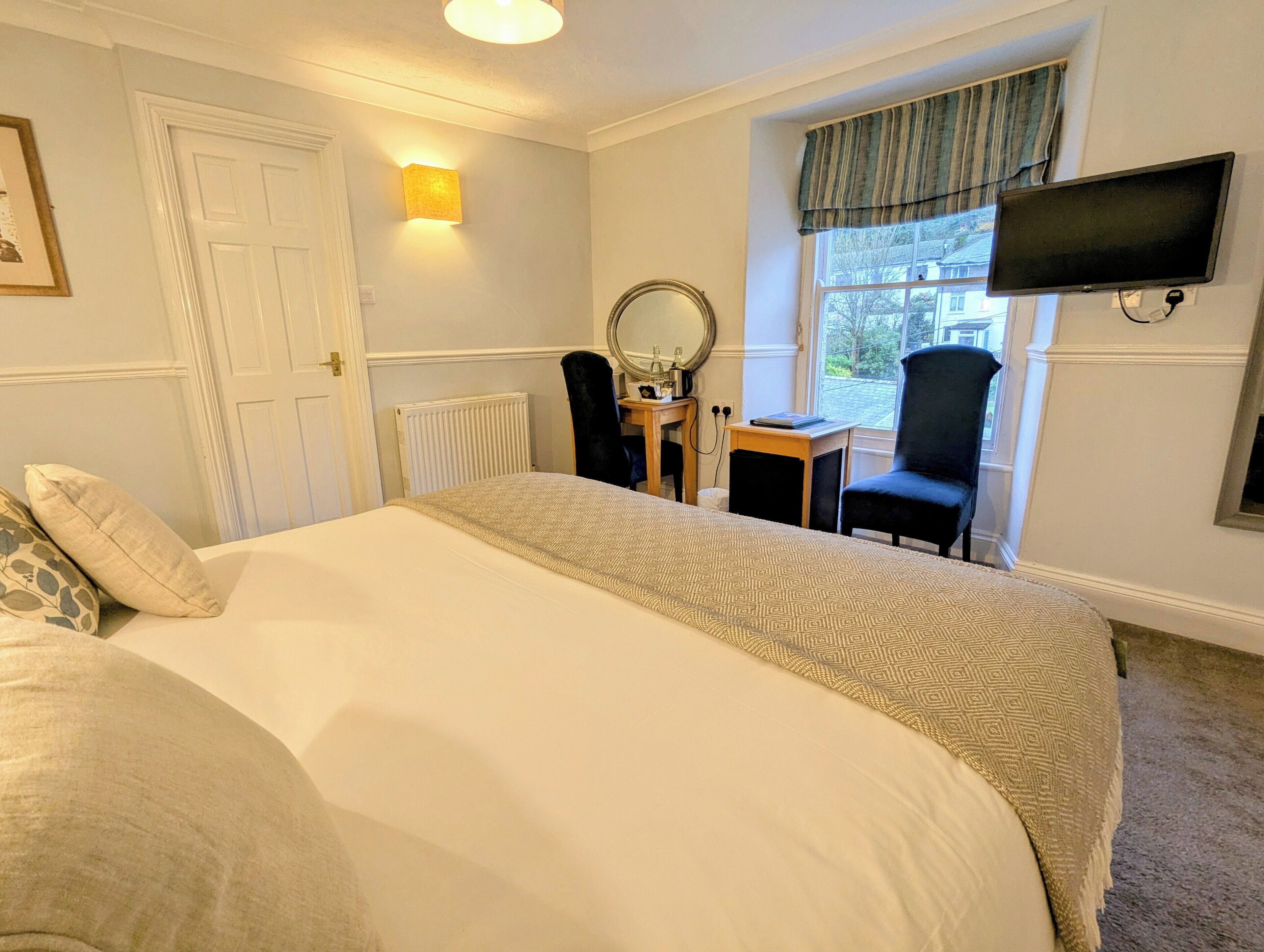 A bedroom with a bed, a desk, a chair, a mirror, a television, and a window with curtains.