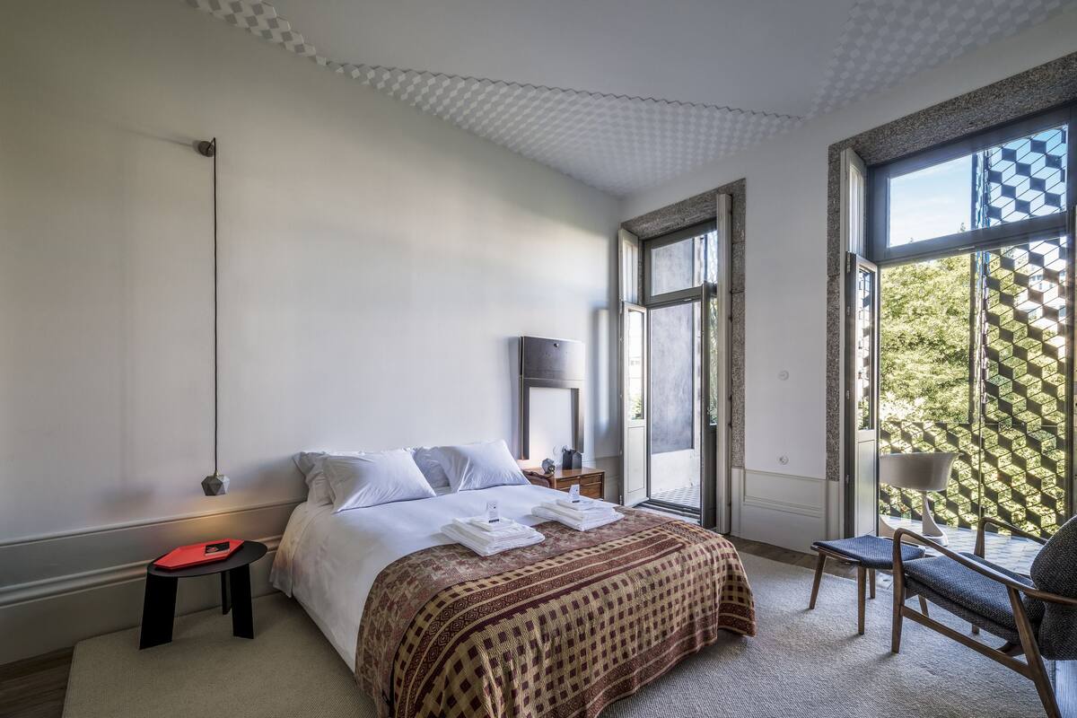 A modern hotel room with a large bed, a small table, a chair, and a large window with a view of greenery.