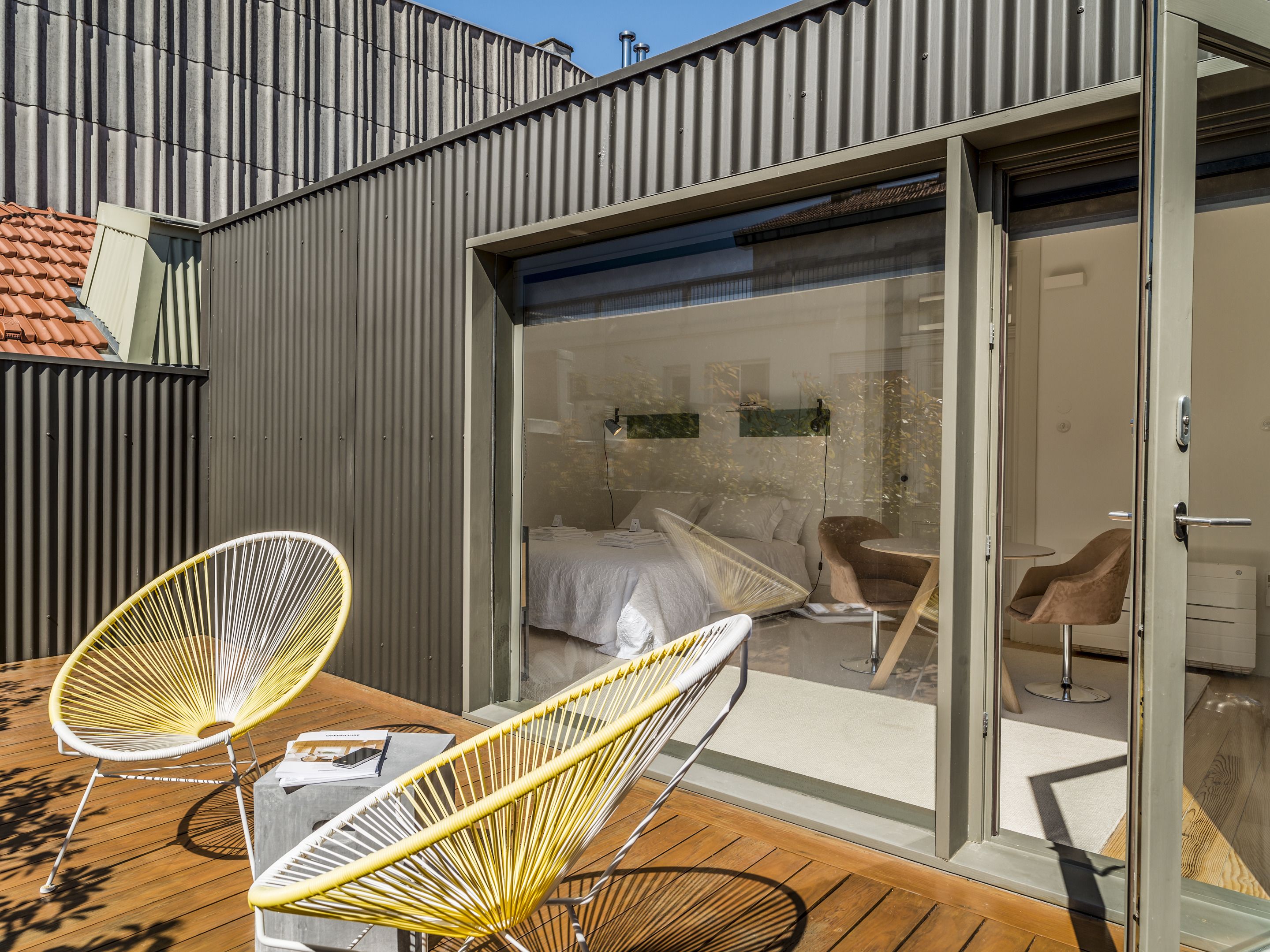 Un patio exterior moderno con dos sillas amarillas, una mesa de madera y un ventanal que da a un dormitorio con una cama, dos sillas y una mesita.
