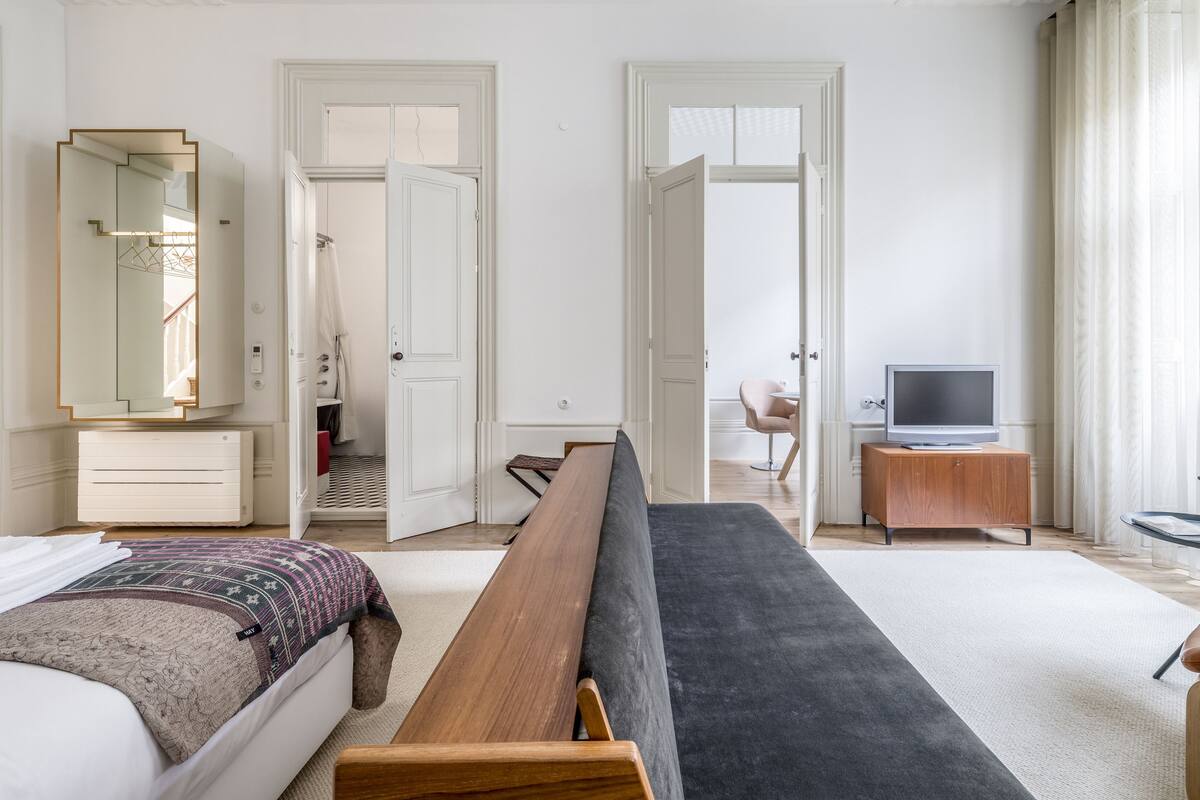 A modern hotel room with a bed, a bench, a TV, and a bathroom visible through an open door.