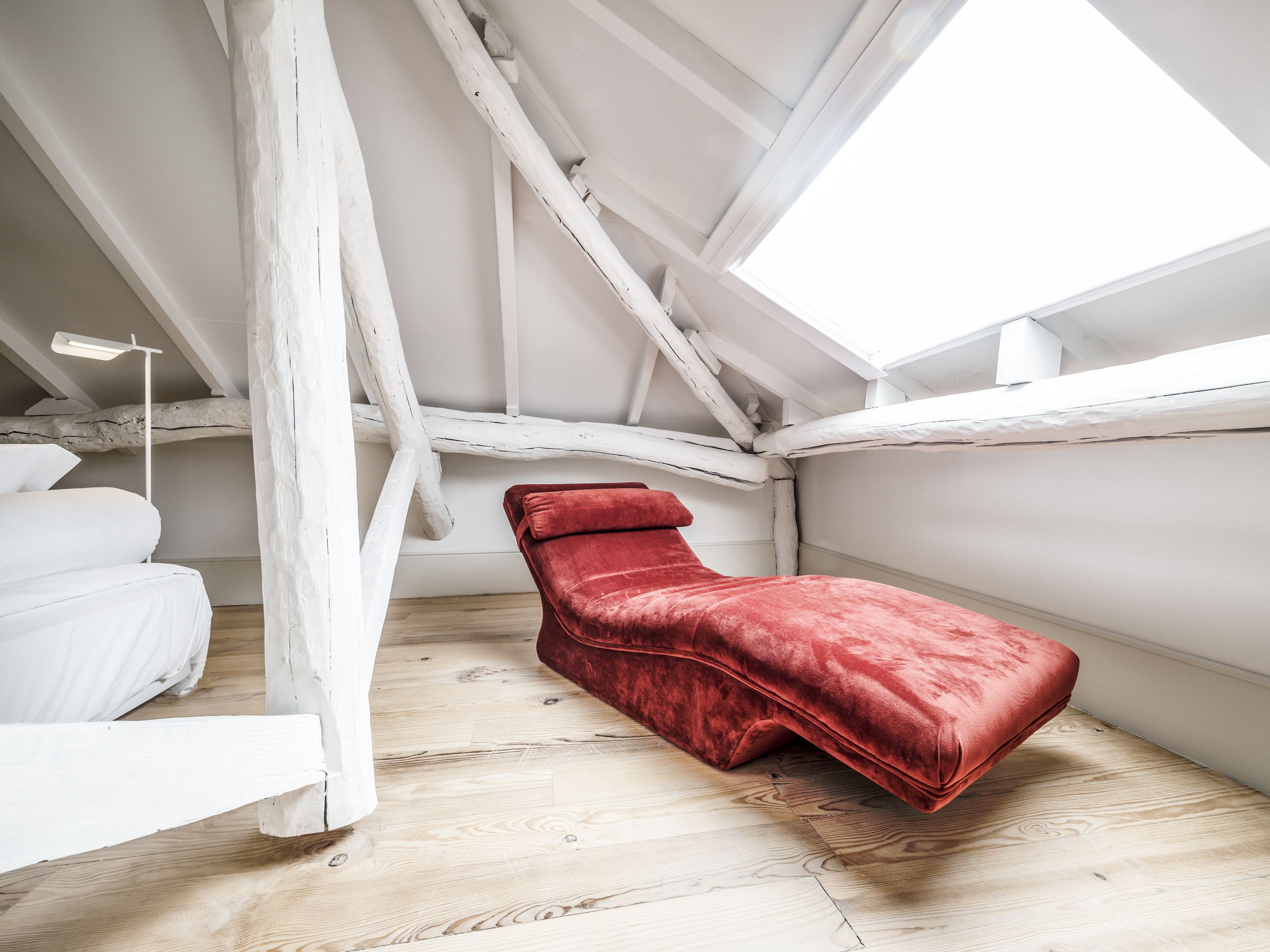 A loft bedroom with a red chaise lounge, white bedding, and a skylight.
