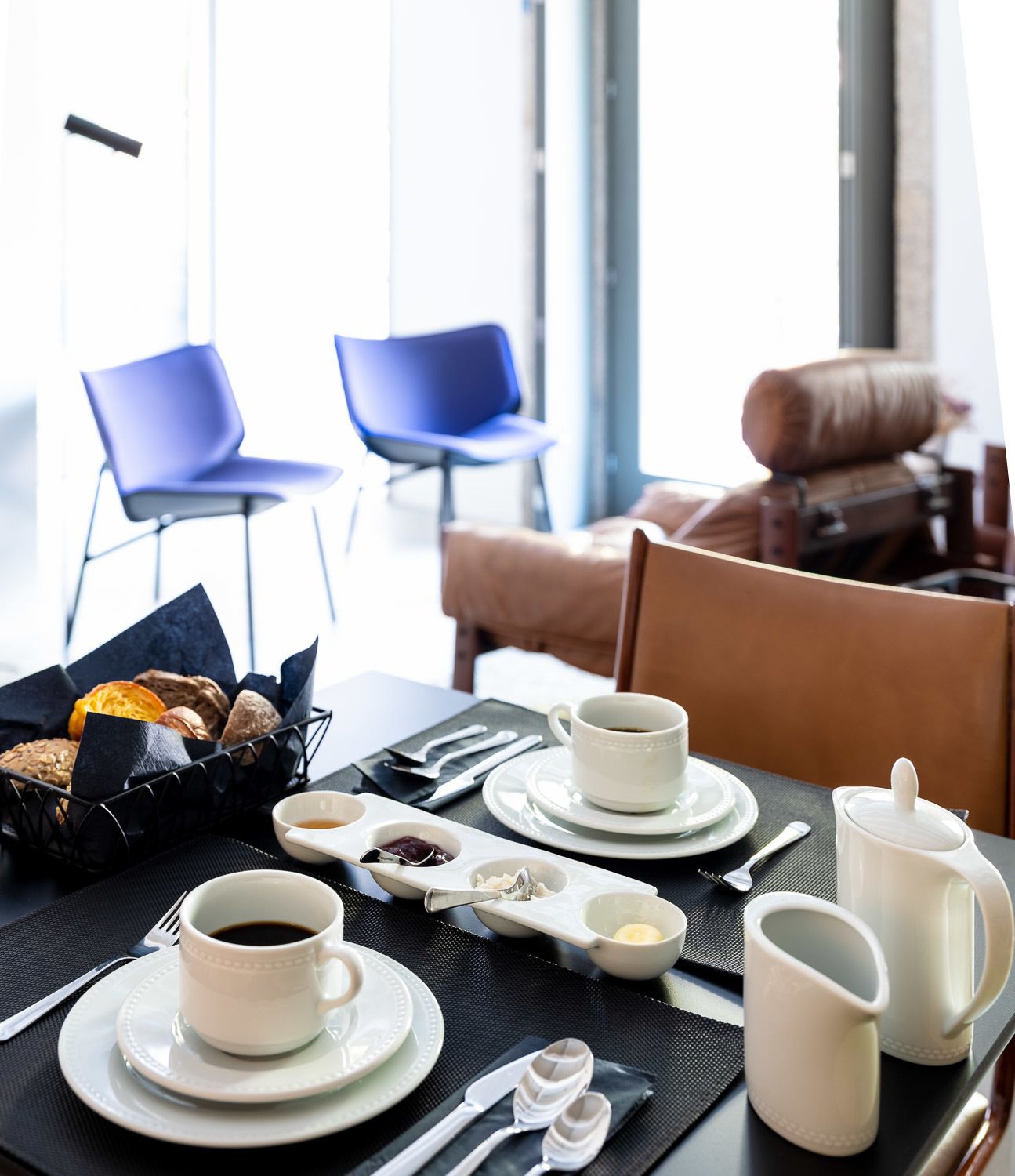 Een ontbijtsetting met een gedekte tafel voor twee personen, inclusief borden, bekers, een mandje met gebak en een koffiepot.
