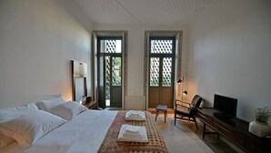 Quarto de hotel com cama, escrivaninha com cadeira, televisão e janela com vista para o verde.