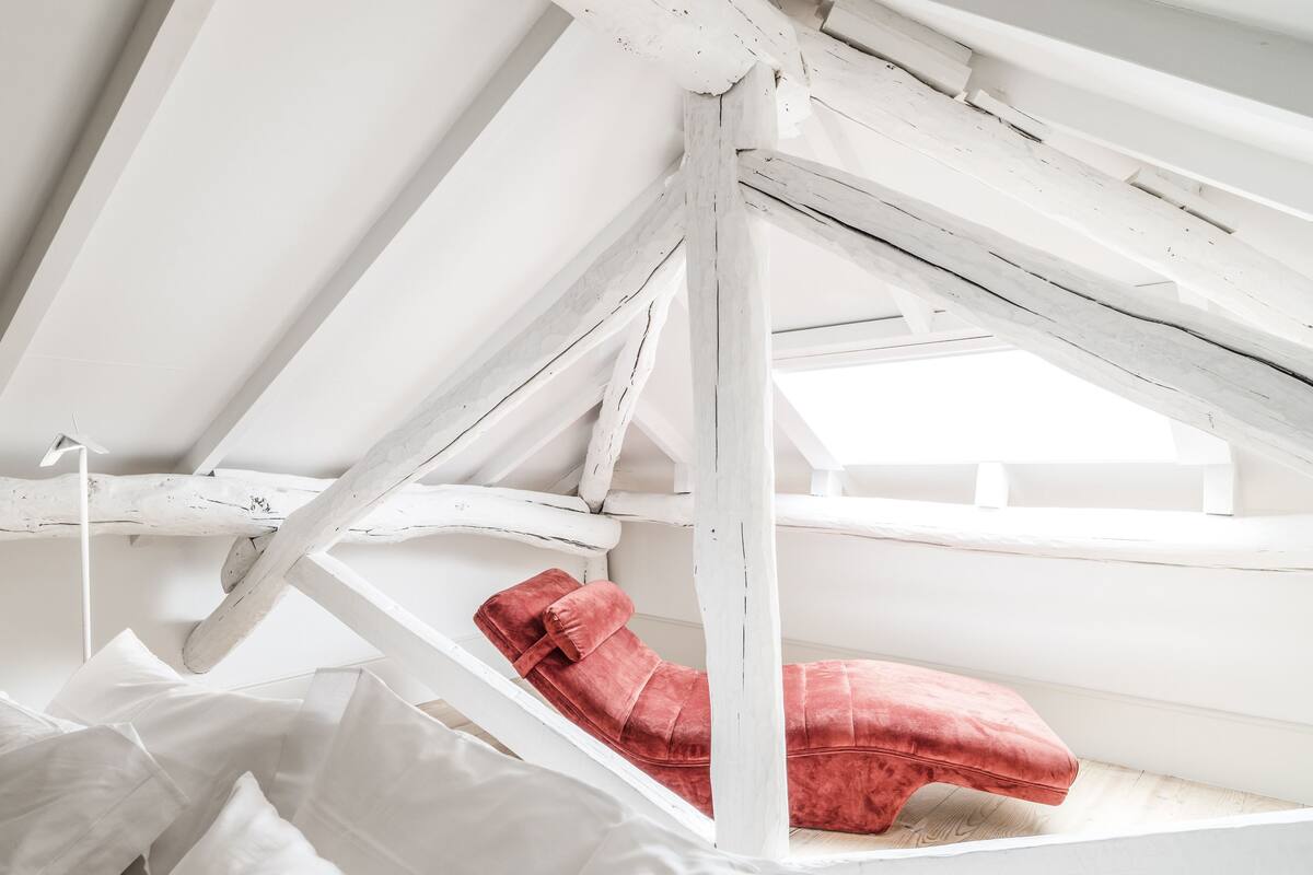 A white, rustic attic bedroom with exposed beams and a red velvet bed.