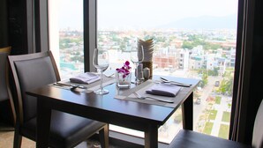 A dining table set with cutlery, a vase with an orchid, and a view of a cityscape through a large window. - Danaciti Hotel (Da Nang)