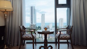 A room with two wooden chairs, a small round table, and a floor lamp. The room has large windows with a view of tall buildings and the sea. - Danaciti Hotel (Da Nang)