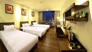 A hotel room with two beds, a desk with a lamp, a small table with a plant, and a view of a cityscape at night. - Danaciti Hotel (Da Nang)