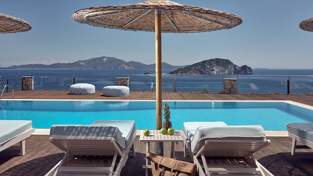 A poolside area with lounge chairs and umbrellas, a wooden deck, and a view of the sea and mountains.
