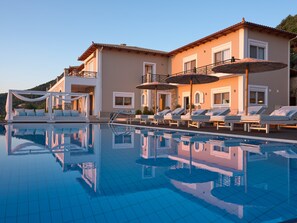 Ein Poolbereich mit Liegestühlen und Sonnenschirmen, das klare Wasser spiegelt die Umgebung wider, und ein Gebäude mit Balkon und Fenstern.
